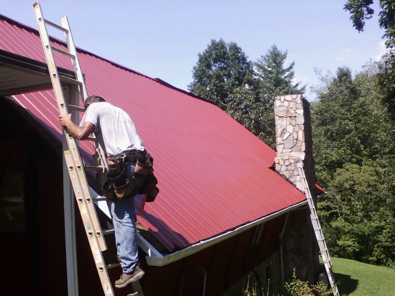 Roofer on a ladder beside a red metal roof and stone chimney — Soffit & Fascia Repair by Roofing Brothers in Loretto